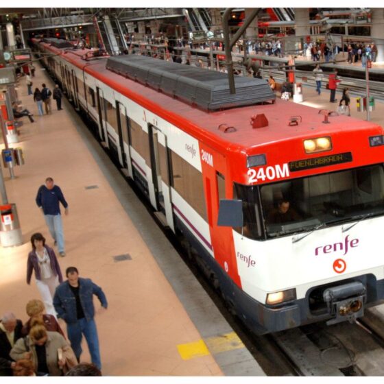 Red and white Renfe commuter train on a platform with several passengers waiting and boarding nearby.