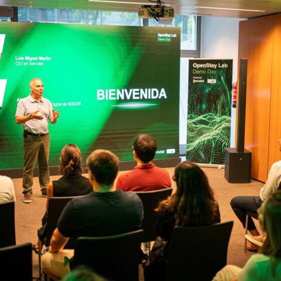 Presenter stands at front of a conference room, delivering a talk in front of a large green screen that reads BIENVENIDA, with headshots on the left and an audience watching.
