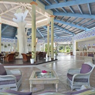 Open-air hotel lobby with wicker chairs around a glass coffee table and a large white tree sculpture at the center, blue-painted ceiling and columns nearby.