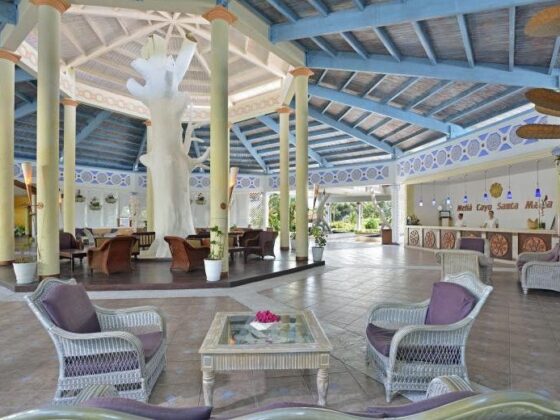 Open-air hotel lobby with wicker chairs around a glass coffee table and a large white tree sculpture at the center, blue-painted ceiling and columns nearby.
