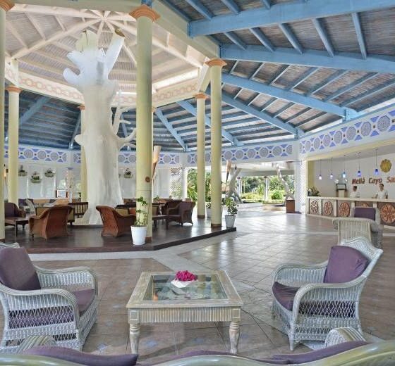 Open-air hotel lobby with wicker chairs around a glass coffee table and a large white tree sculpture at the center, blue-painted ceiling and columns nearby.