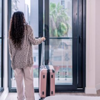 Back view of a woman in a knit sweater and light pants, standing by a glass door with a pink rolling suitcase beside her in a bright interior.