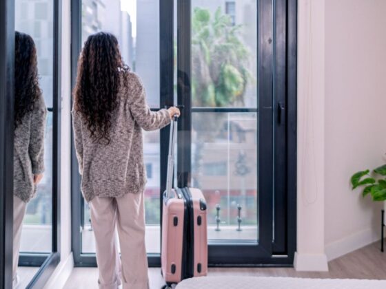 Back view of a woman in a knit sweater and light pants, standing by a glass door with a pink rolling suitcase beside her in a bright interior.