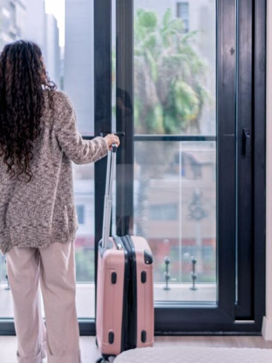 Back view of a woman in a knit sweater and light pants, standing by a glass door with a pink rolling suitcase beside her in a bright interior.
