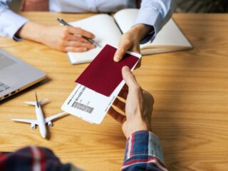 Hands exchanging a burgundy passport and boarding pass over a wooden table, with a laptop and small airplane model nearby.