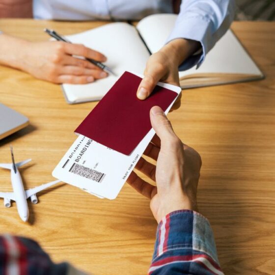Hands exchanging a burgundy passport and boarding pass over a wooden table, with a laptop and small airplane model nearby.