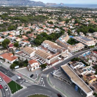 Aerial view of a coastal town showing a large roundabout, beige residential buildings, and tree-covered hills in the distance.