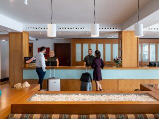 Hotel lobby reception with guests and staff at a wooden check-in desk, luggage nearby, and glass-front cabinets behind.