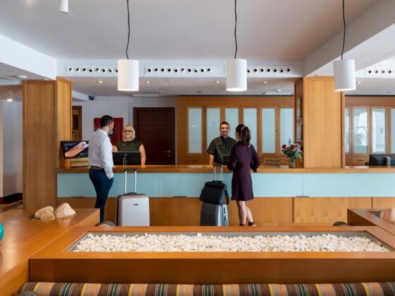 Hotel lobby reception with guests and staff at a wooden check-in desk, luggage nearby, and glass-front cabinets behind.