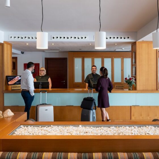 Hotel lobby reception with guests and staff at a wooden check-in desk, luggage nearby, and glass-front cabinets behind.