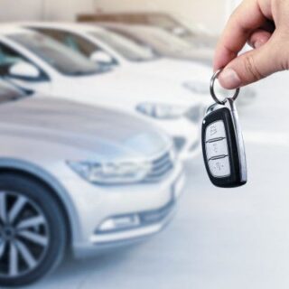 Person holding a car key fob up in front of parked cars, ready to unlock a vehicle at a dealership or showroom.