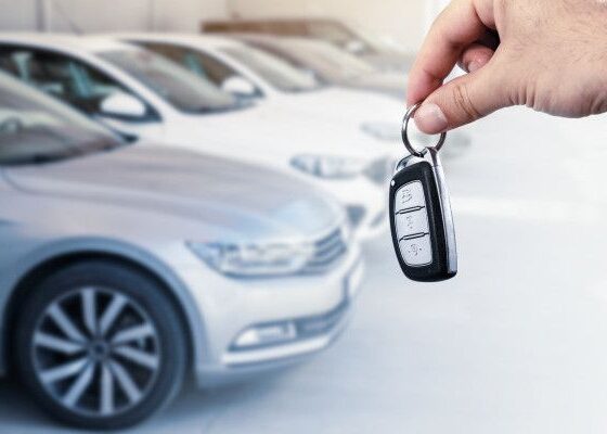 Person holding a car key fob up in front of parked cars, ready to unlock a vehicle at a dealership or showroom.