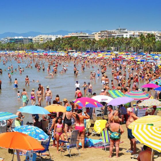 Busy sunny beach packed with people under colorful umbrellas; calm shallow water and high-rise buildings in the background.
