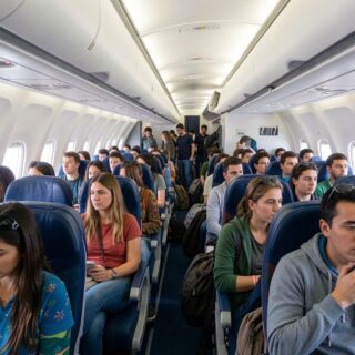 Crowded airplane cabin with passengers seated in blue seats along the aisle under overhead bins.