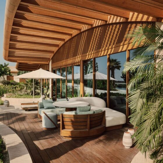 Modern outdoor lounge on a wooden deck with curved white sofa, teal cushions, and a round table under a white umbrella by glass windows.