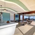 Bright beachfront cafe interior with curved green ceiling panels, white bar, and seating by large windows overlooking the outside sea view.