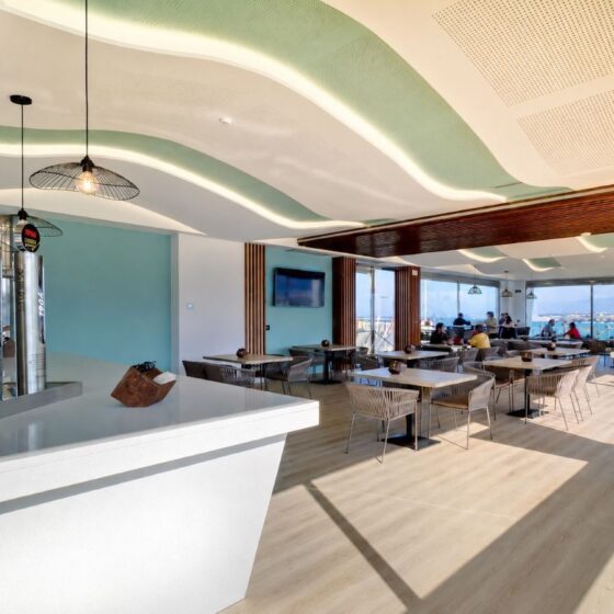 Bright beachfront cafe interior with curved green ceiling panels, white bar, and seating by large windows overlooking the outside sea view.