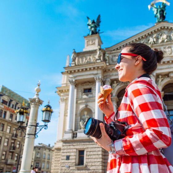 Woman in a red plaid shirt with sunglasses holds ice cream and a camera, in front of an ornate historic building on a sunny day