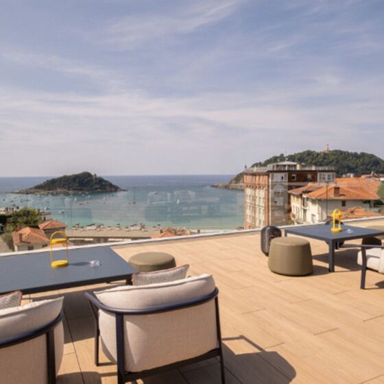 Rooftop terrace with dark tables and beige seating overlooking a calm sea and small island beyond, under a blue sky.