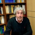 Middle-aged man with gray curly hair and stubble seated at a desk in a library, wearing a pink collared shirt under a dark sweater, smiling slightly.