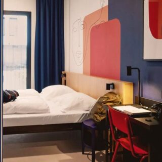 Hotel room with a white bed, wooden headboard, blue walls, and a red chair at a desk by the window.