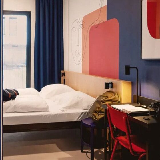 Hotel room with a white bed, wooden headboard, blue walls, and a red chair at a desk by the window.