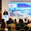 Speaker at a podium giving a tourism marketing presentation; large screen shows a beach photo of Las Palmas.