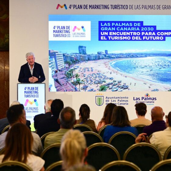 Speaker at a podium giving a tourism marketing presentation; large screen shows a beach photo of Las Palmas.