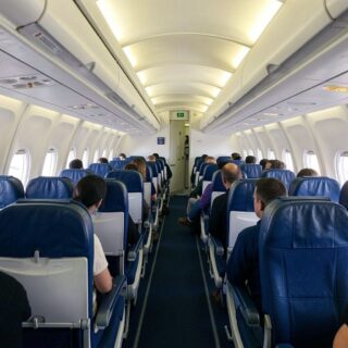 Inside a commercial airplane cabin: passengers seated in blue seats along a central aisle, windows on both sides, view toward the forward door.