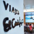 Travel agency interior with a wall sign reading 'VIAJES' and staff at desks near a large beach photo mural in the background.