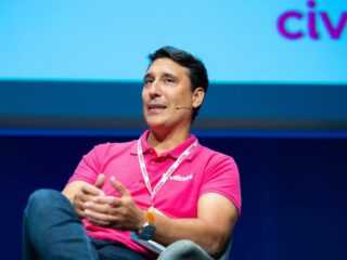 Man in a bright pink polo speaking on stage with a headset mic and conference lanyard.