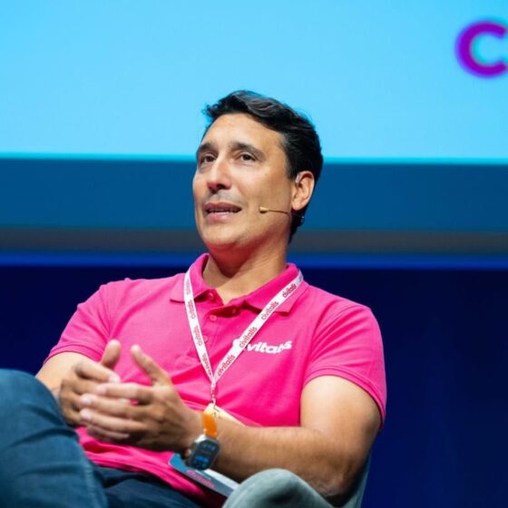 Man in a bright pink polo speaking on stage with a headset mic and conference lanyard.