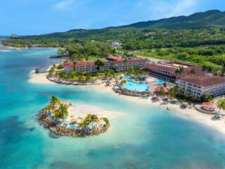 A tropical beachfront resort with white buildings, a central blue pool, palm trees, and a curved sandy beach by turquoise water.