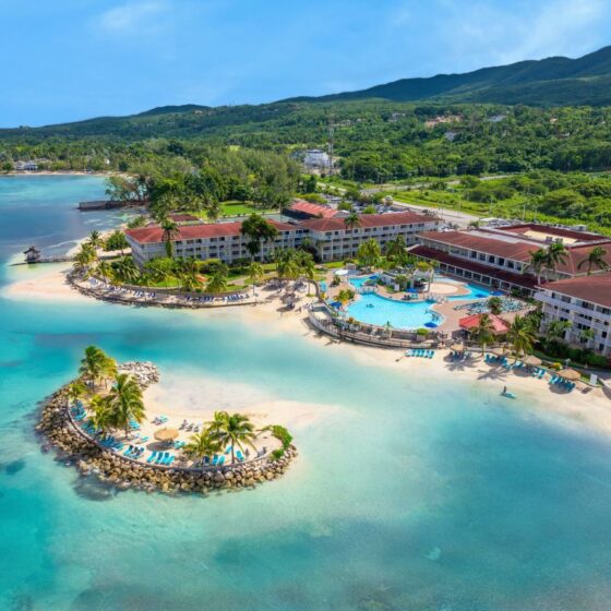 A tropical beachfront resort with white buildings, a central blue pool, palm trees, and a curved sandy beach by turquoise water.