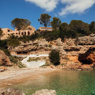 Cliffside cove with clear turquoise water, rocky beach, and a tan arched building atop the cliff among pine trees.