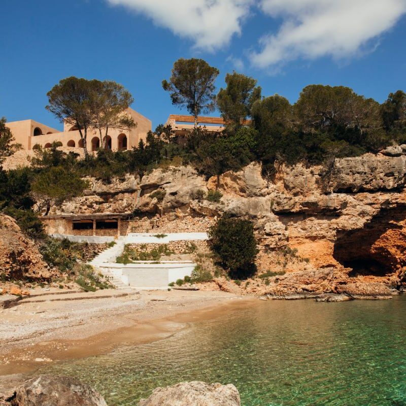 Cliffside cove with clear turquoise water, rocky beach, and a tan arched building atop the cliff among pine trees.