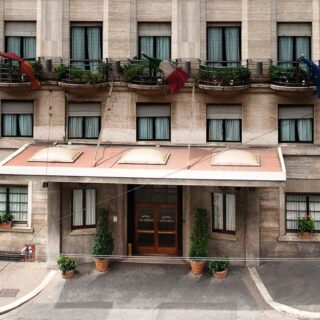 Facade of a beige hotel with a covered entrance, potted plants, and balconies along the upper floors; three flags flutter above the awning.