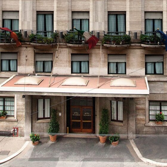 Facade of a beige hotel with a covered entrance, potted plants, and balconies along the upper floors; three flags flutter above the awning.