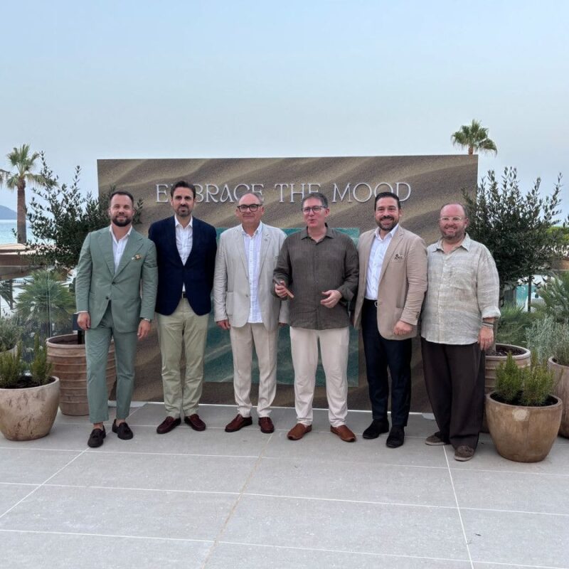 Six men in smart casuals stand on a terrace in front of a sign reading 'Embrace the Mood' with the sea behind them.