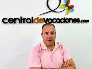 Man in a pink polo shirt stands with arms crossed in front of a wall displaying the centraldevacaciones.com logo with a colorful flower graphic.