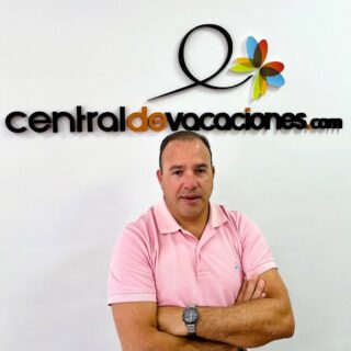 Man in a pink polo shirt stands with arms crossed in front of a wall displaying the centraldevacaciones.com logo with a colorful flower graphic.