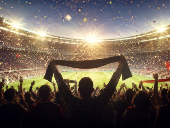 Crowd of stadium spectators celebrating, a fan in the foreground raises a scarf above their head as lights blaze overhead.