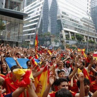 Crowd of Spain soccer fans waving red and yellow flags in a city plaza, raising hands in celebration near modern glass buildings.