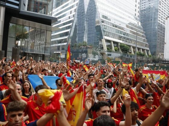 Crowd of Spain soccer fans waving red and yellow flags in a city plaza, raising hands in celebration near modern glass buildings.