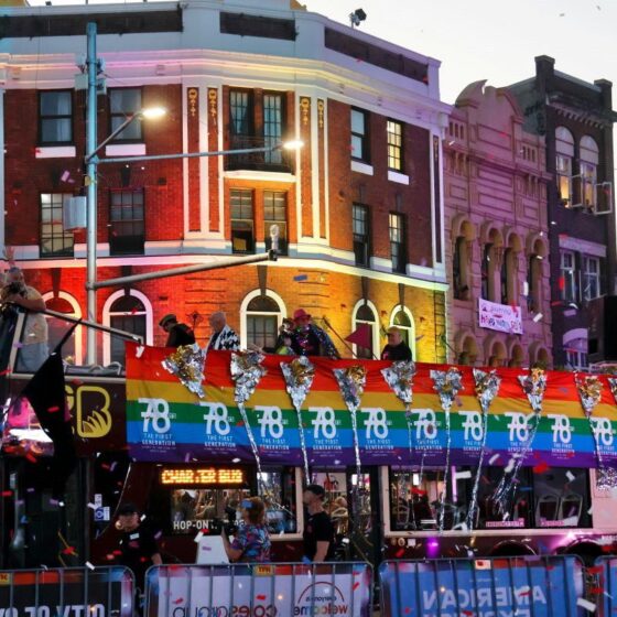 Colorful Pride parade float on a city street with rainbow banners, silver confetti, and people on the upper deck celebrating.