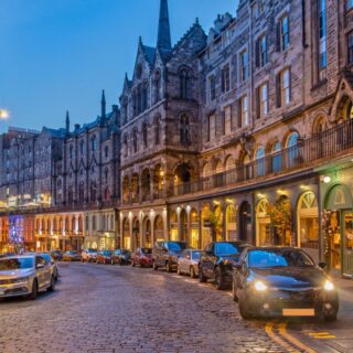 Cobblestone street lined with historic stone buildings and arched arcades at dusk, cars parked along the curb.
