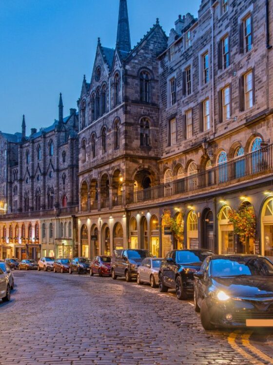 Cobblestone street lined with historic stone buildings and arched arcades at dusk, cars parked along the curb.