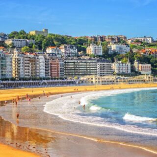 Sunny beach with golden sand and blue-green waves, lined by colorful apartment buildings and green hills in the background.