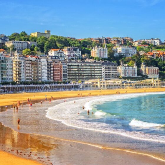 Sunny beach with golden sand and blue-green waves, lined by colorful apartment buildings and green hills in the background.