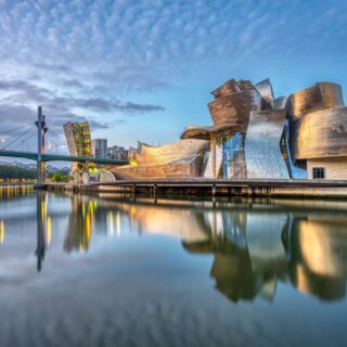 The Guggenheim Museum Bilbao's sculptural metallic façade reflects in calm river water at dusk, with a bridge to the left and city lights in the background.